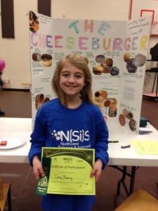 Cora Baerg, a grade 3 student at Uplands Elementary School, proudly wears one of the N(Si)S T-shirts and displays her ribbon and certificate, which acknowledge the hard work she put into researching the rate of decay of cheeseburgers made with and without preservatives. Nice job, Cora!